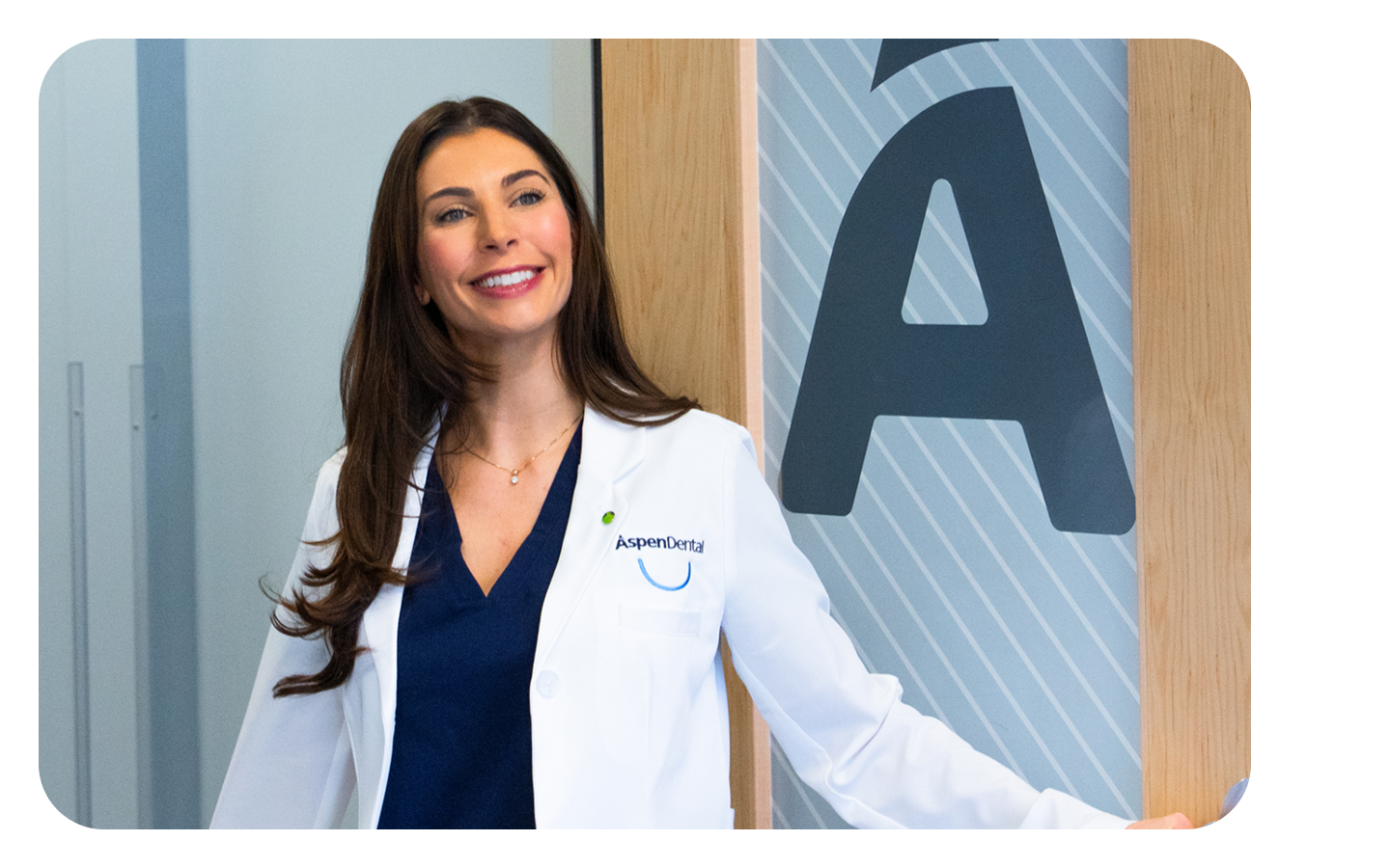 Female dentist in a white coat stands smiling at the entrance of an Aspen Dental location, highlighting the welcoming atmosphere of the office.