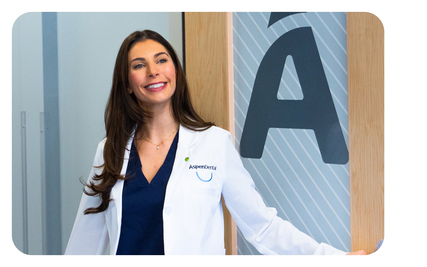 Female dentist in a white coat stands smiling at the entrance of an Aspen Dental location, highlighting the welcoming atmosphere of the office.
