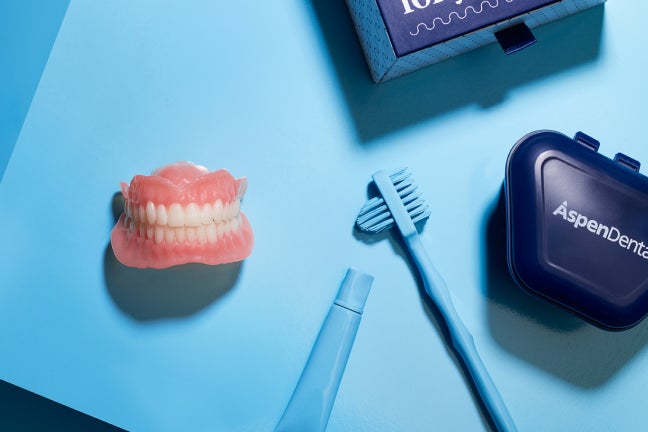 A flat-lay image of a denture care kit, including a full denture model, cleaning tools, and a storage case, displayed on a vibrant blue background for Aspen Dental.