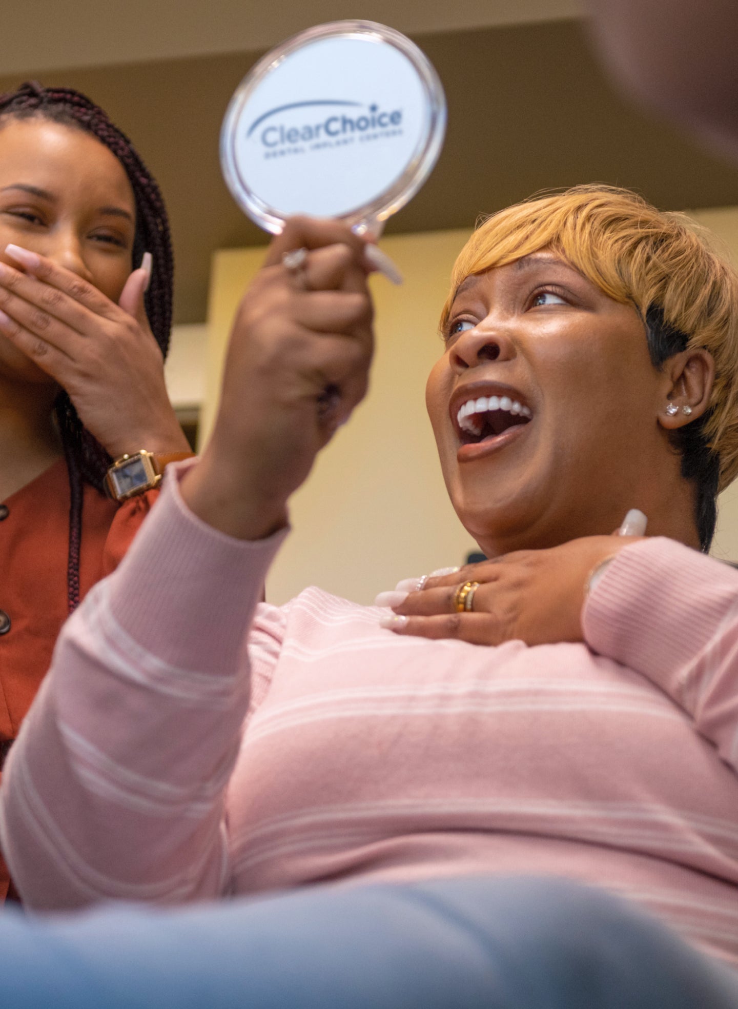 A happy ClearChoice patient holding a mirror and smiling joyfully while a friend smiles in the background, showcasing a successful dental transformation.