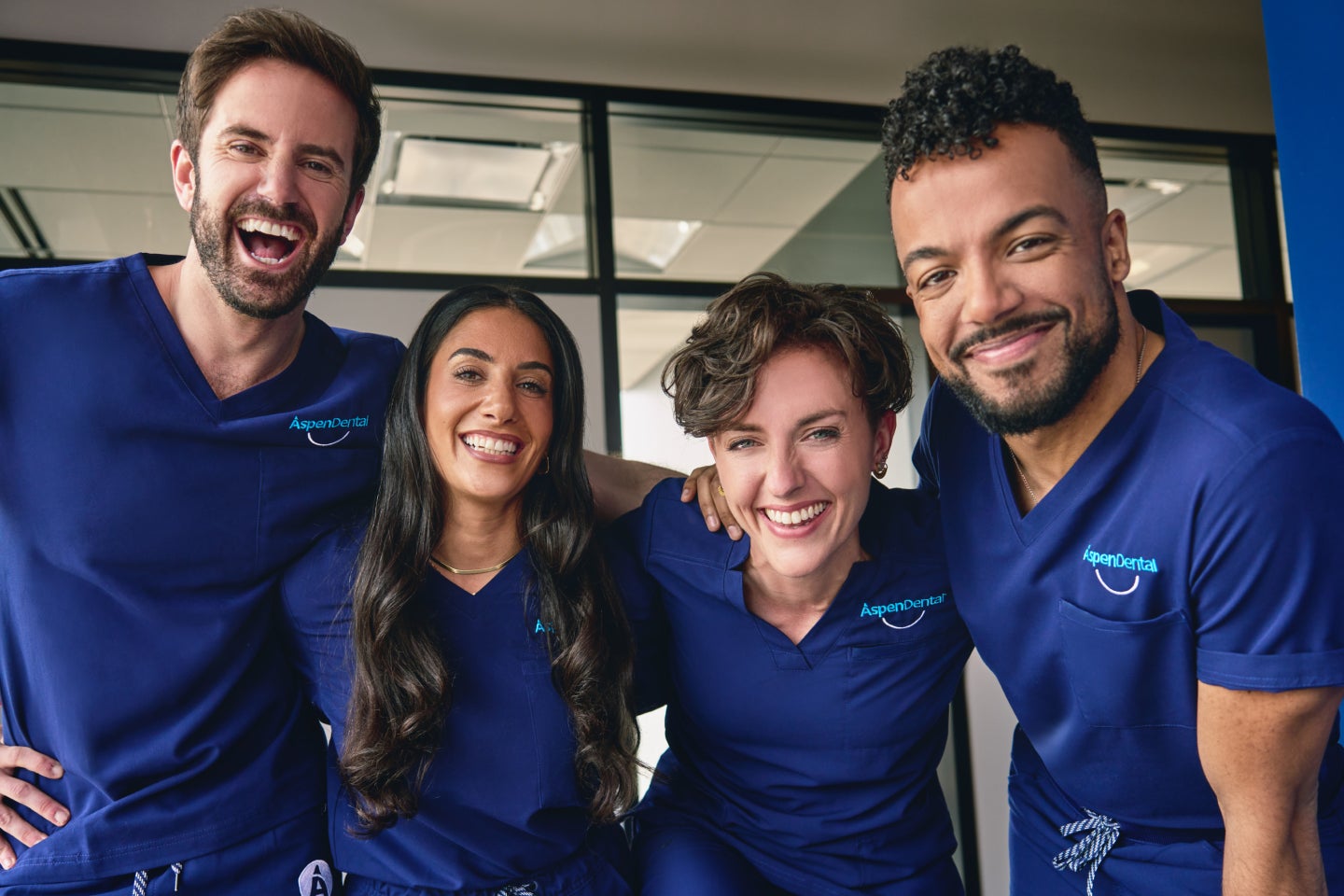 A group of Aspen Dental professionals smiling confidently in their scrubs, standing together in a modern office setting, reflecting teamwork, expertise, and patient-centered care.