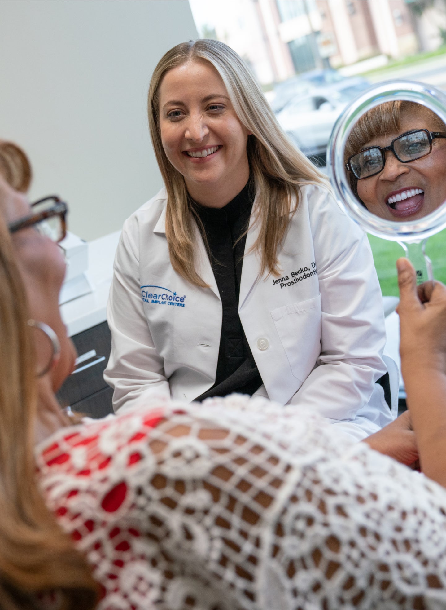 A ClearChoice prosthodontist in a white coat smiling while consulting with a patient. The patient, wearing a red and white lace top, holds a mirror and admires her new smile. The discussion highlights the differences between dental implants and veneers for tooth restoration.
