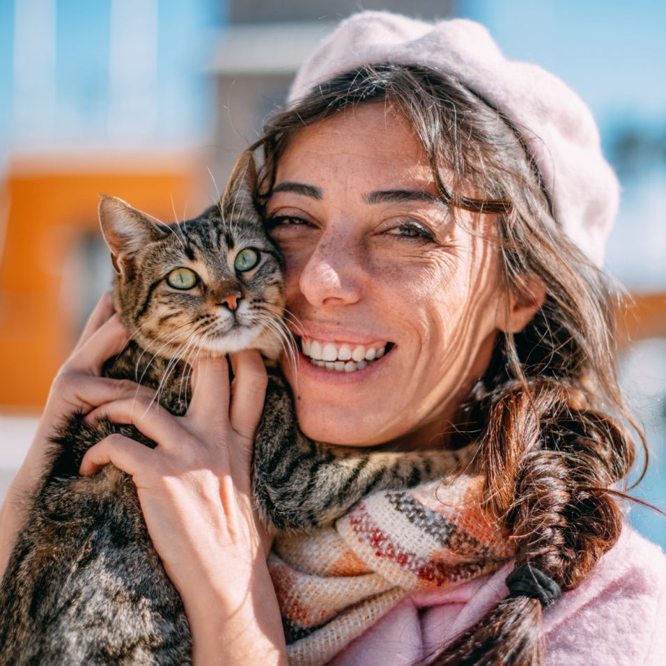 A brunette woman smiles while holding a brown tabby cat outside on a sunny day