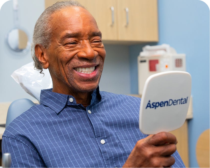 Smiling male patient holds a handheld mirror with the Aspen Dental logo, representing satisfaction with general dental services.