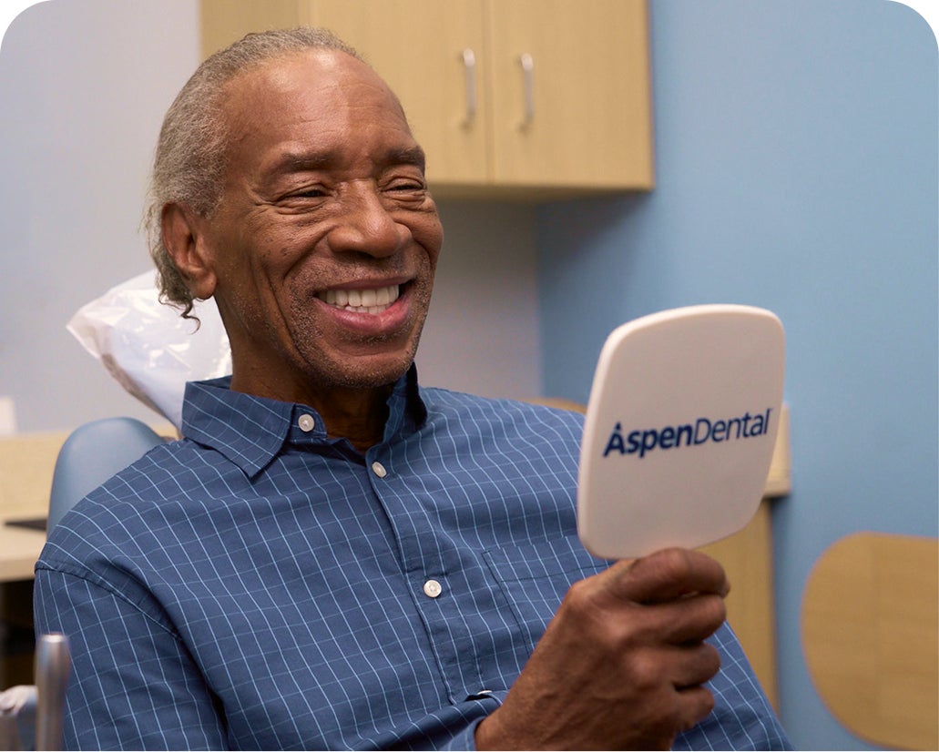 Older man smiles while examining his dentures in an Aspen Dental mirror, representing industry-leading denture warranties.