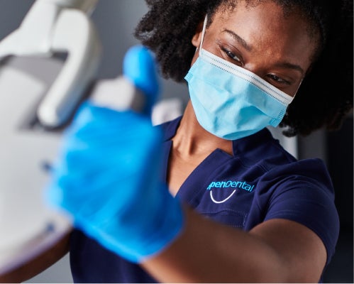 A dental assistant wearing a mask and gloves gives a thumbs-up while preparing dental equipment, representing Aspen Dental's commitment to professional and friendly care.