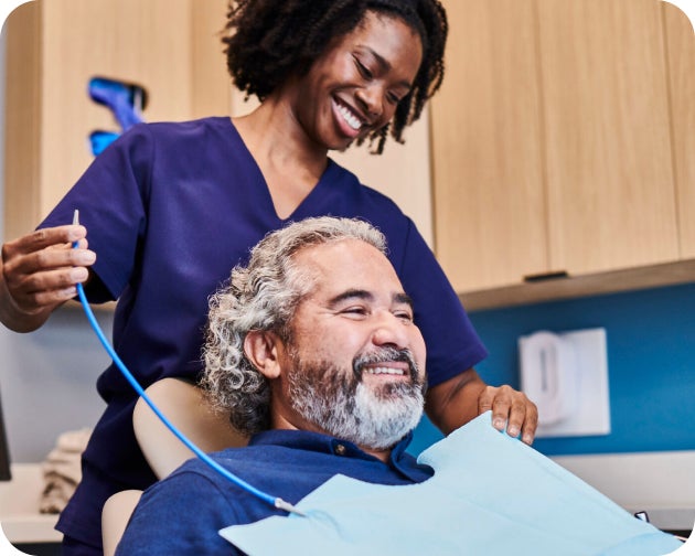 A smiling patient with gray hair and a beard sits in a dental chair during a routine dental checkup, assisted by a friendly dental professional in a purple uniform at an Aspen Dental clinic.
