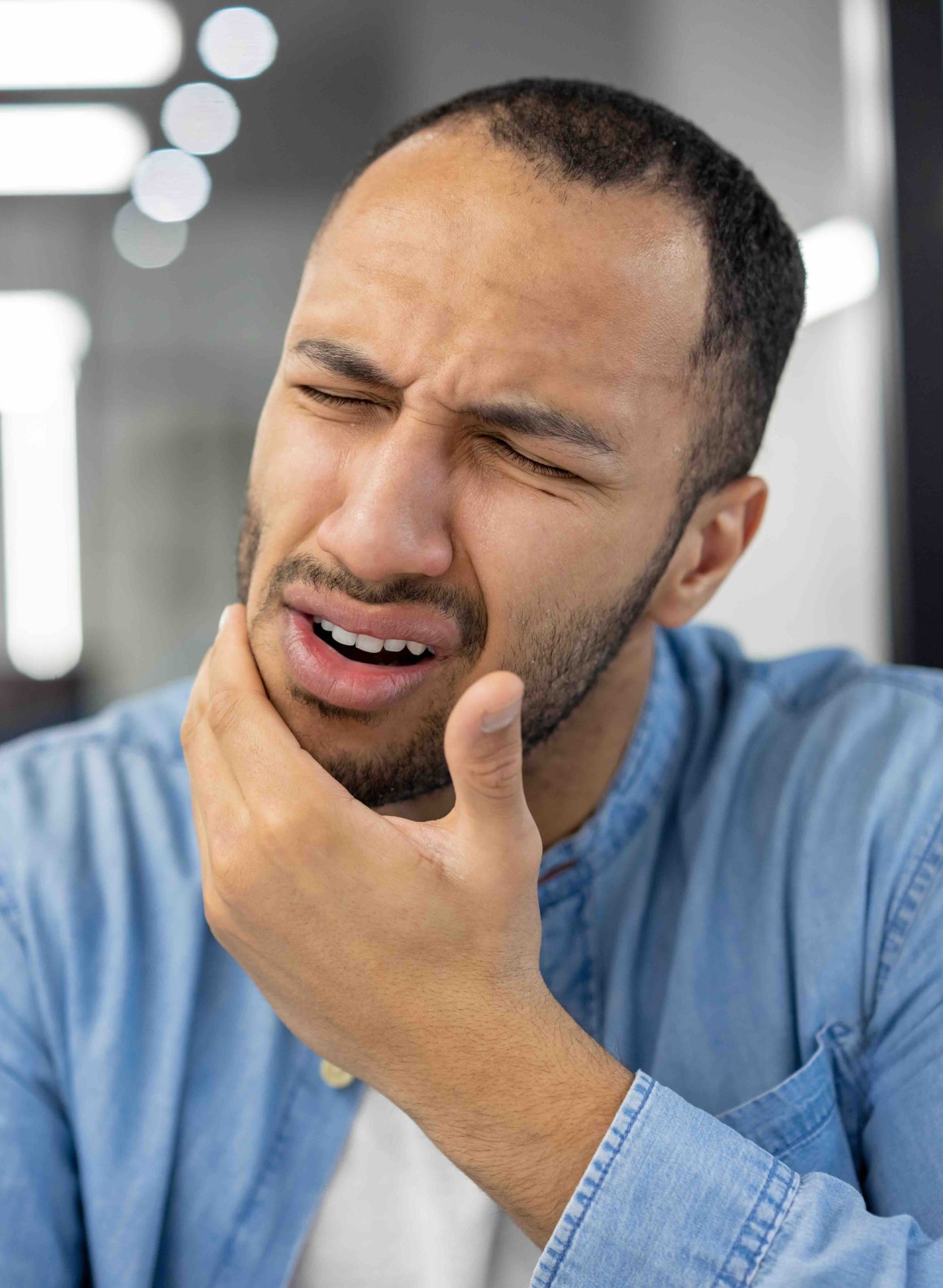 A man wearing a denim shirt holding his jaw with a pained expression, indicating toothache or dental discomfort, possibly due to oral health issues.