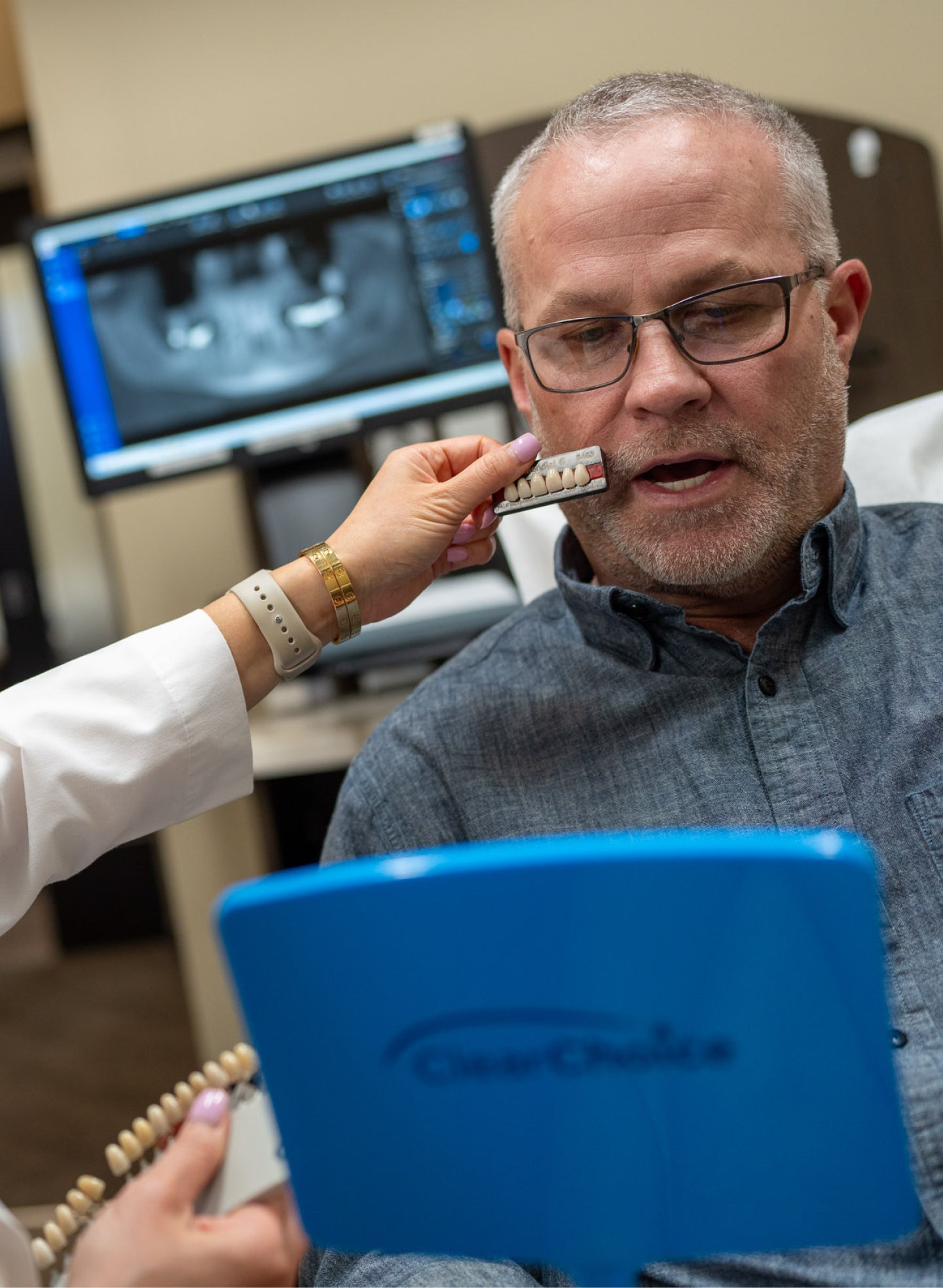 A middle-aged man in a ClearChoice dental clinic is undergoing a dental implant consultation. A dental professional is holding a shade guide to his teeth while he looks into a ClearChoice-branded mirror. A computer screen in the background displays a dental X-ray, showing implant placement and abutment options.