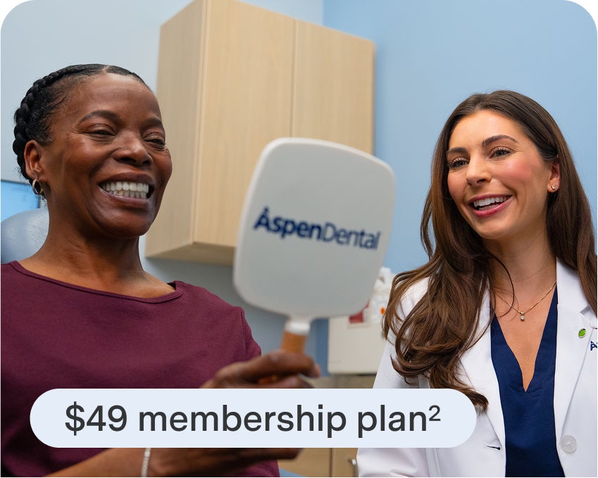 Smiling patient admiring her teeth in an Aspen Dental mirror while talking with a friendly dental professional, showing the $49 membership plan.