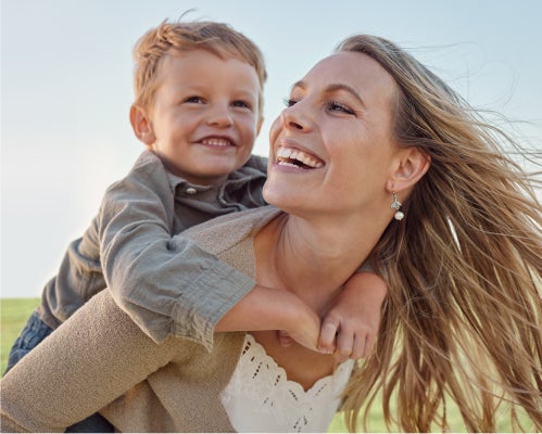 A joyful mother giving her son a piggyback ride in a sunny outdoor setting, symbolizing the confidence and happiness brought by Aspen Dental's comprehensive care.