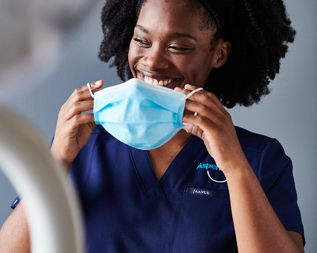 A female dental professional with curly hair smiles while putting on a face mask, preparing for a patient appointment. Dental experts at Aspen Dental ensure a safe and professional experience for optimal oral health care.