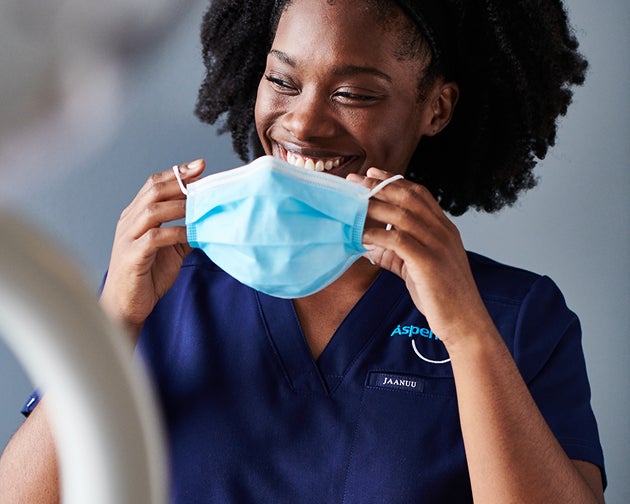 A female dental professional with curly hair smiles while putting on a face mask, preparing for a patient appointment. Dental experts at Aspen Dental ensure a safe and professional experience for optimal oral health care.