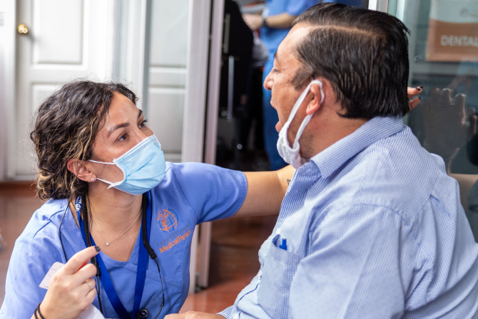 A compassionate Aspen Dental team member wearing a blue uniform and mask provides assistance to a seated patient during the Healthy Mouth Movement initiative.