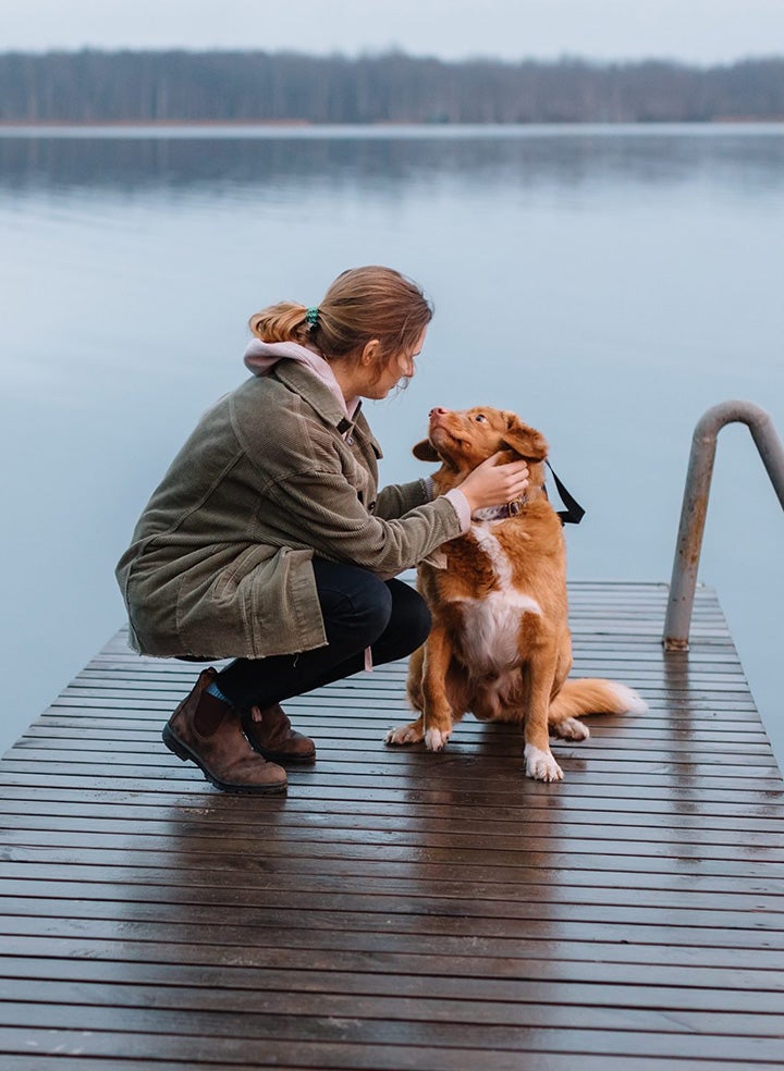 A woman bending down to scratch her dog's neck. They are standing on a dock overlooking a vast lake.