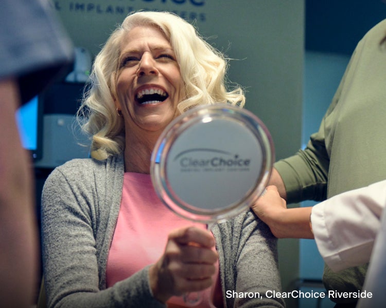 Sharon, a ClearChoice dental implant patient, looking into a hand mirror. She is smiling and admiring the dental implant work from ClearChoice.