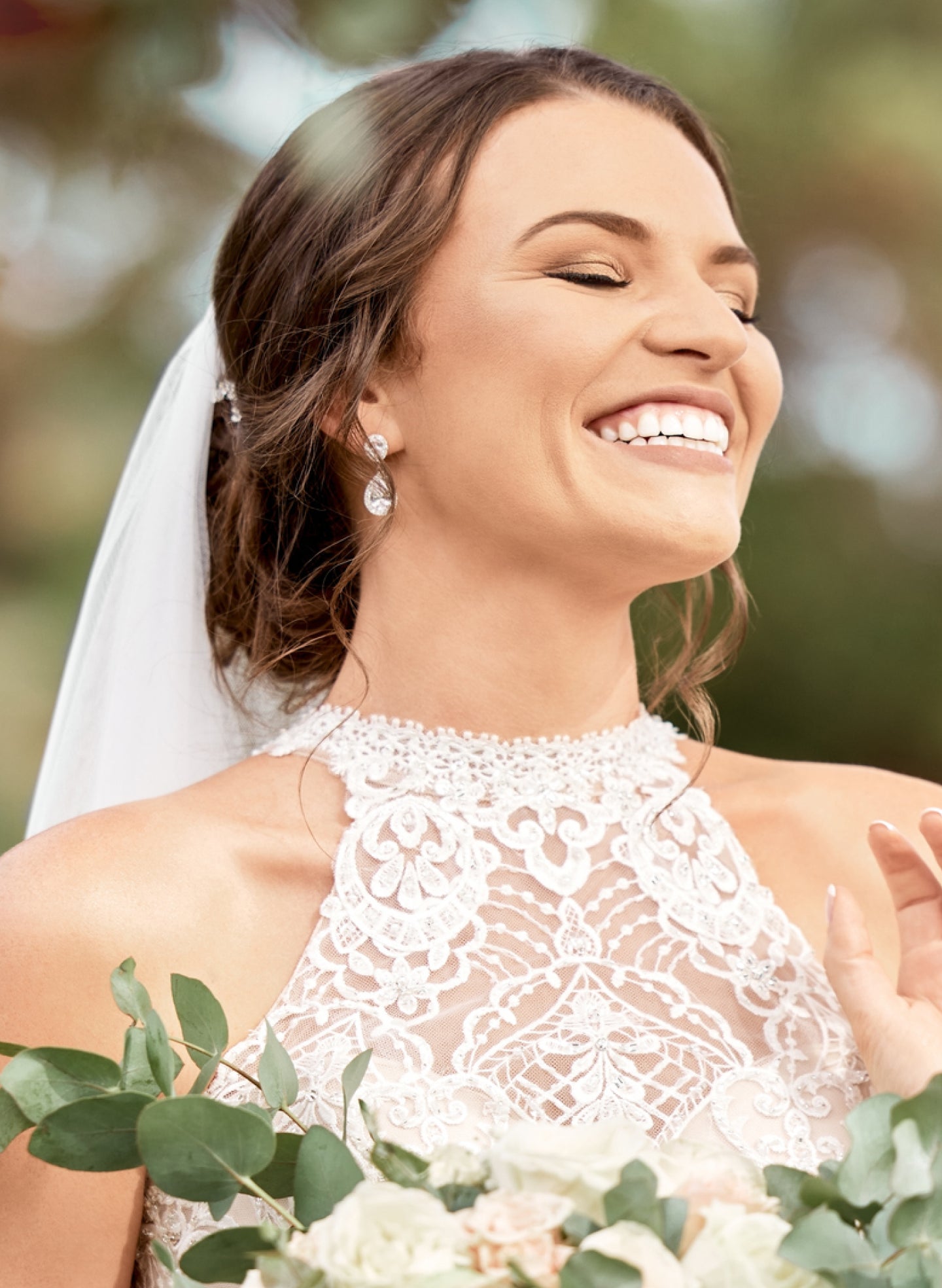 A bride in a lace wedding dress and veil, smiling with eyes closed, holding a bouquet of white flowers.