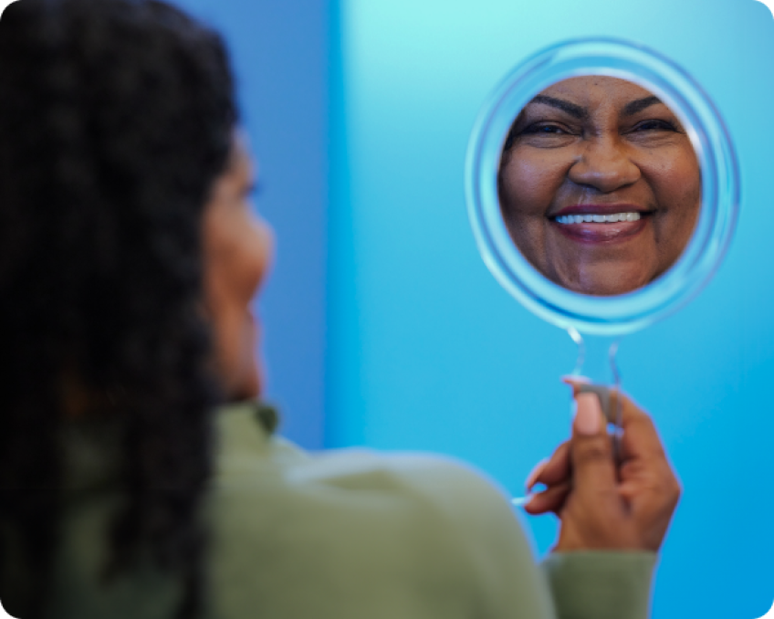 A happy Aspen Dental patient admiring their smile in a handheld mirror after successful wisdom tooth extraction and dental care, with a bright blue background.