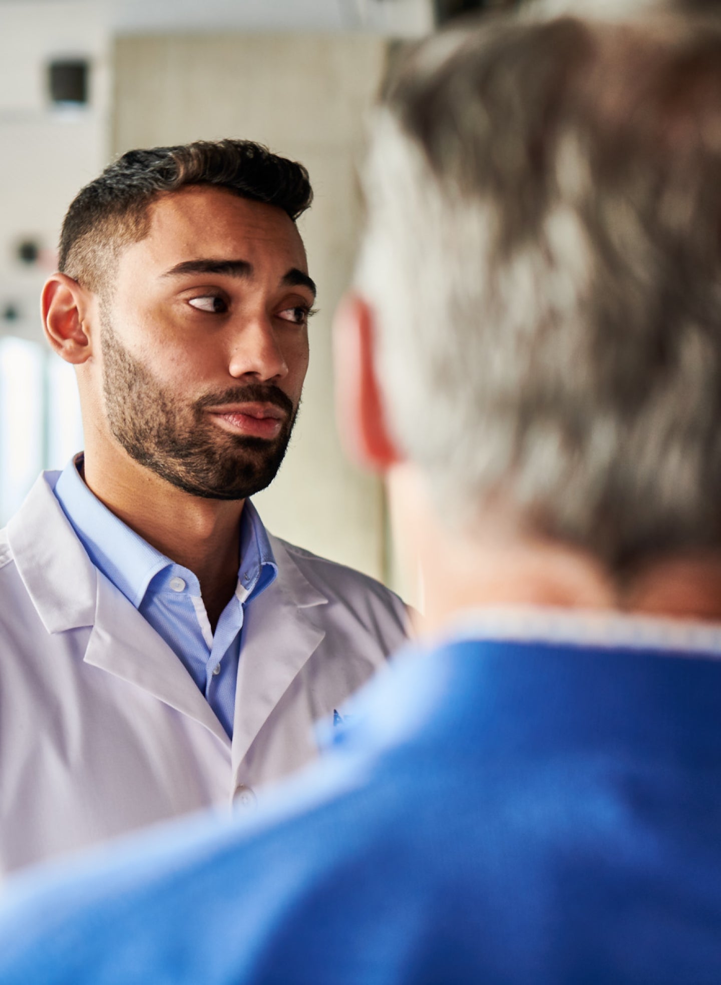 A man in a white lab coat talks to another man with grey hair dressed in blue.