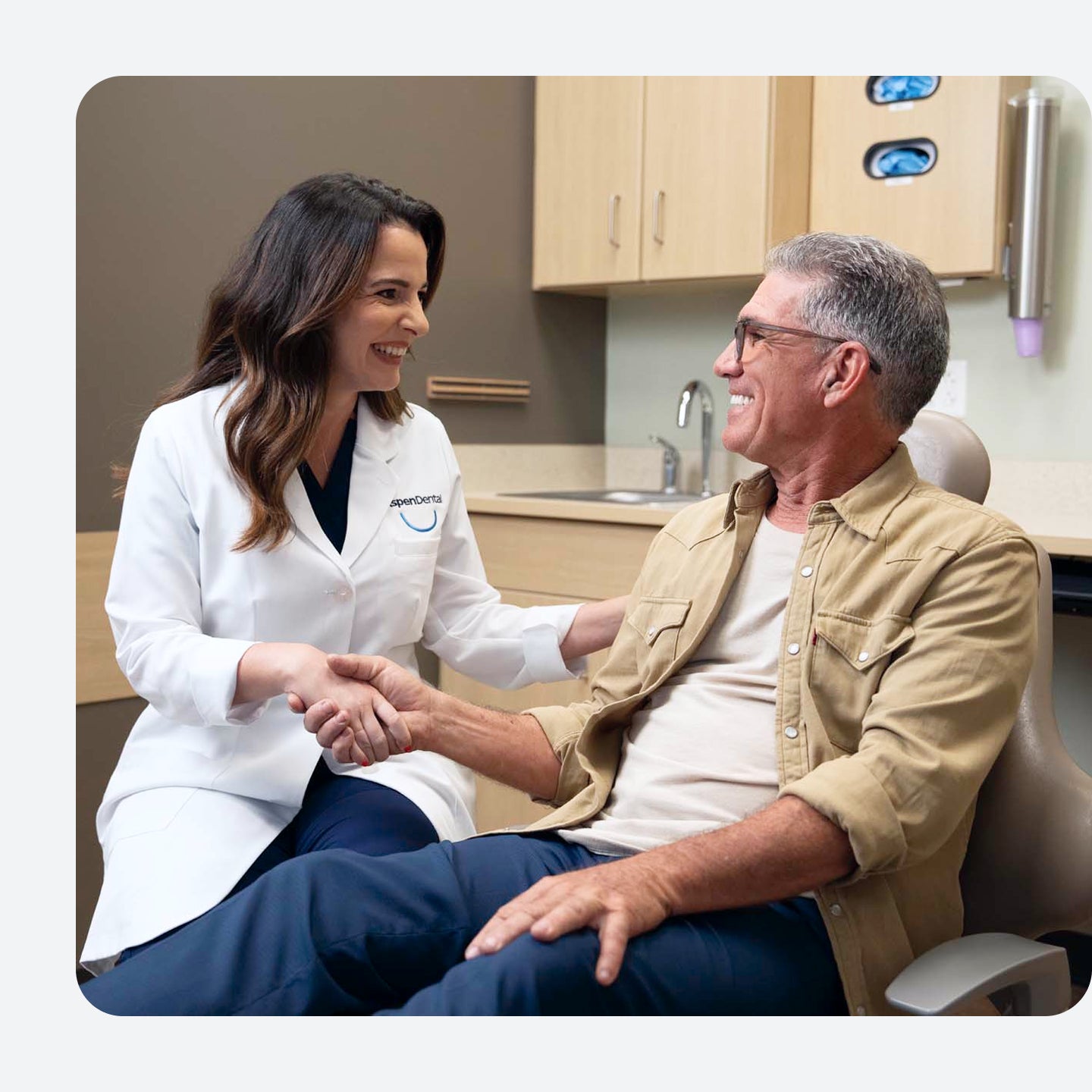 Una profesional dental de Aspen Dental sonriente con bata blanca estrecha cálidamente la mano de un paciente masculino mayor en una sala de examen dental. Parecen tener una conversación amistosa y tranquilizadora, lo que sugiere una experiencia dental positiva.