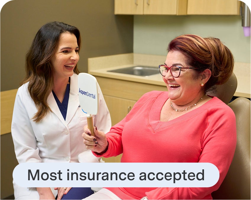 Aspen Dental patient admires her smile in a mirror after learning her dental insurance is accepted, with a dental provider sitting beside her.
