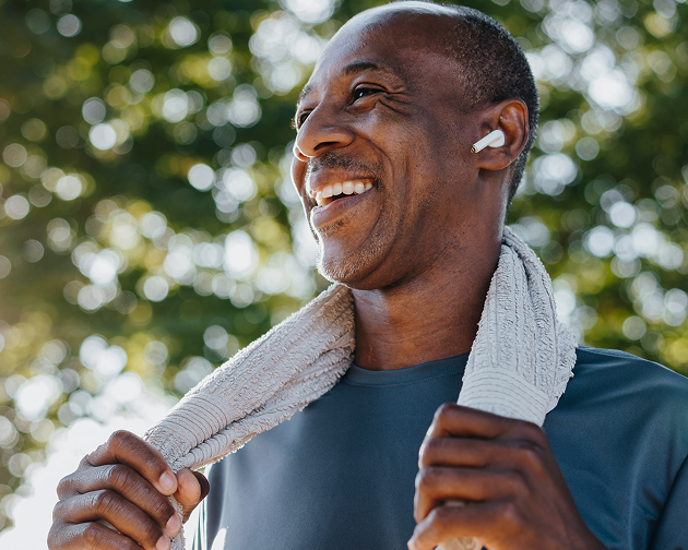 A fit, smiling man with earbuds and a towel around his neck enjoys the fresh air after a workout. Maintaining a healthy lifestyle, including exercise and oral care, contributes to overall well-being. Aspen Dental supports holistic health for a confident smile.