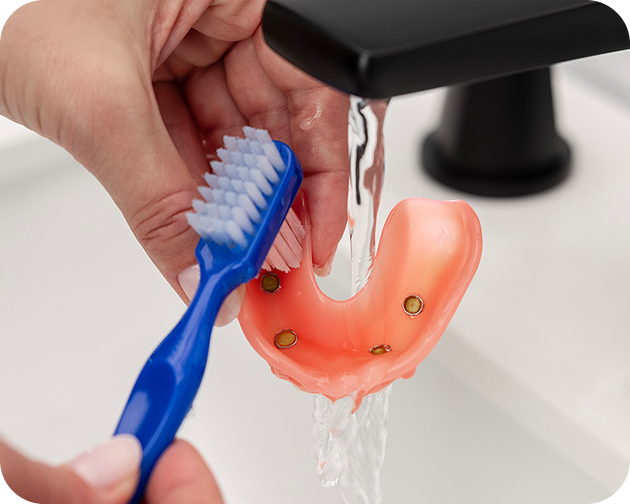 Cleaning an implant denture under running water using a blue toothbrush – Aspen Dental oral hygiene care.