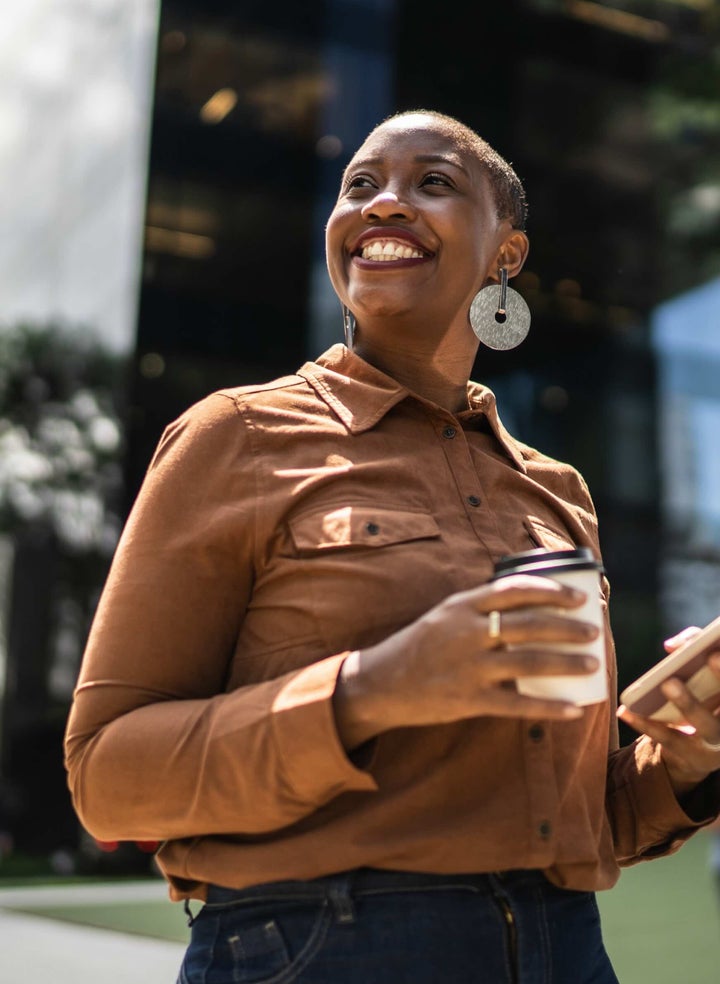 A smiling woman holding a coffee cup and phone outdoors, showcasing her bright, white teeth after professional teeth whitening treatment, emphasizing confidence and a radiant smile.