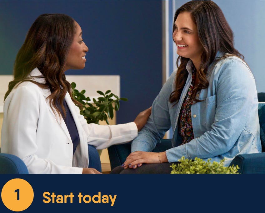 Smiling Motto dental professional in a white coat engaging warmly with a patient seated beside her, creating a comforting atmosphere in a modern Motto dental office setting with greenery in the background.