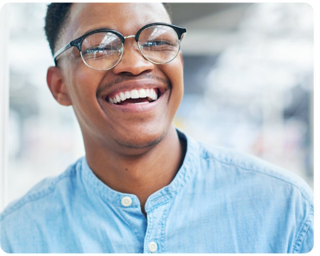 A cheerful man wearing round glasses and a light blue casual shirt, smiling confidently in a bright indoor setting, representing Aspen Dental's expertise in titanium dental implants and their welcoming, professional care approach.