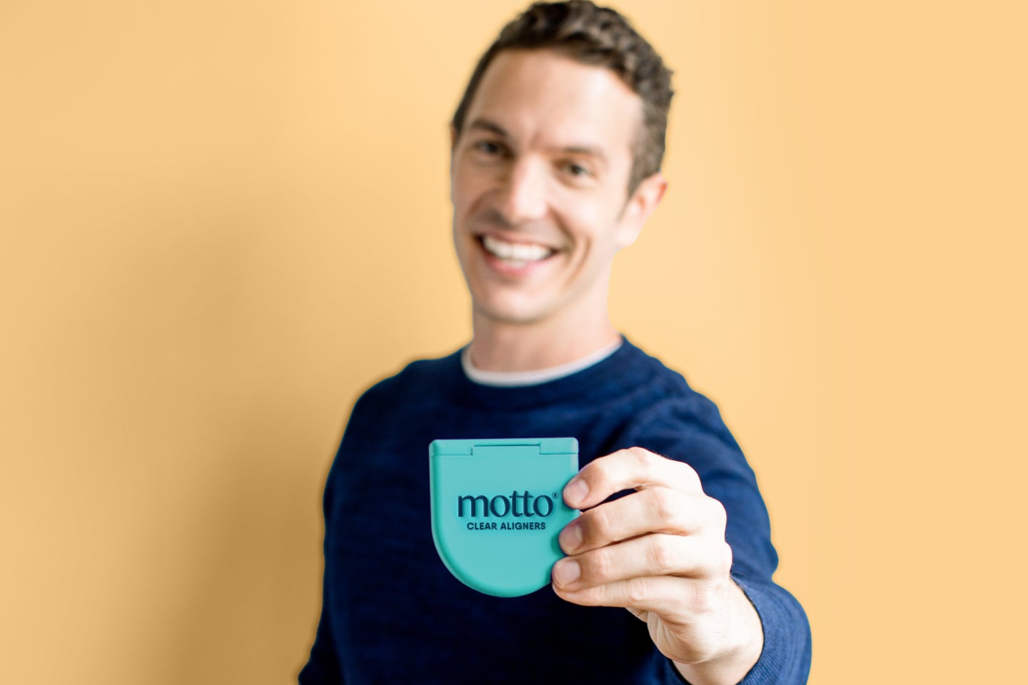  A man smiling confidently while holding a Motto clear aligner case in focus, set against a soft yellow background, showcasing the ease and convenience of Motto aligners.
