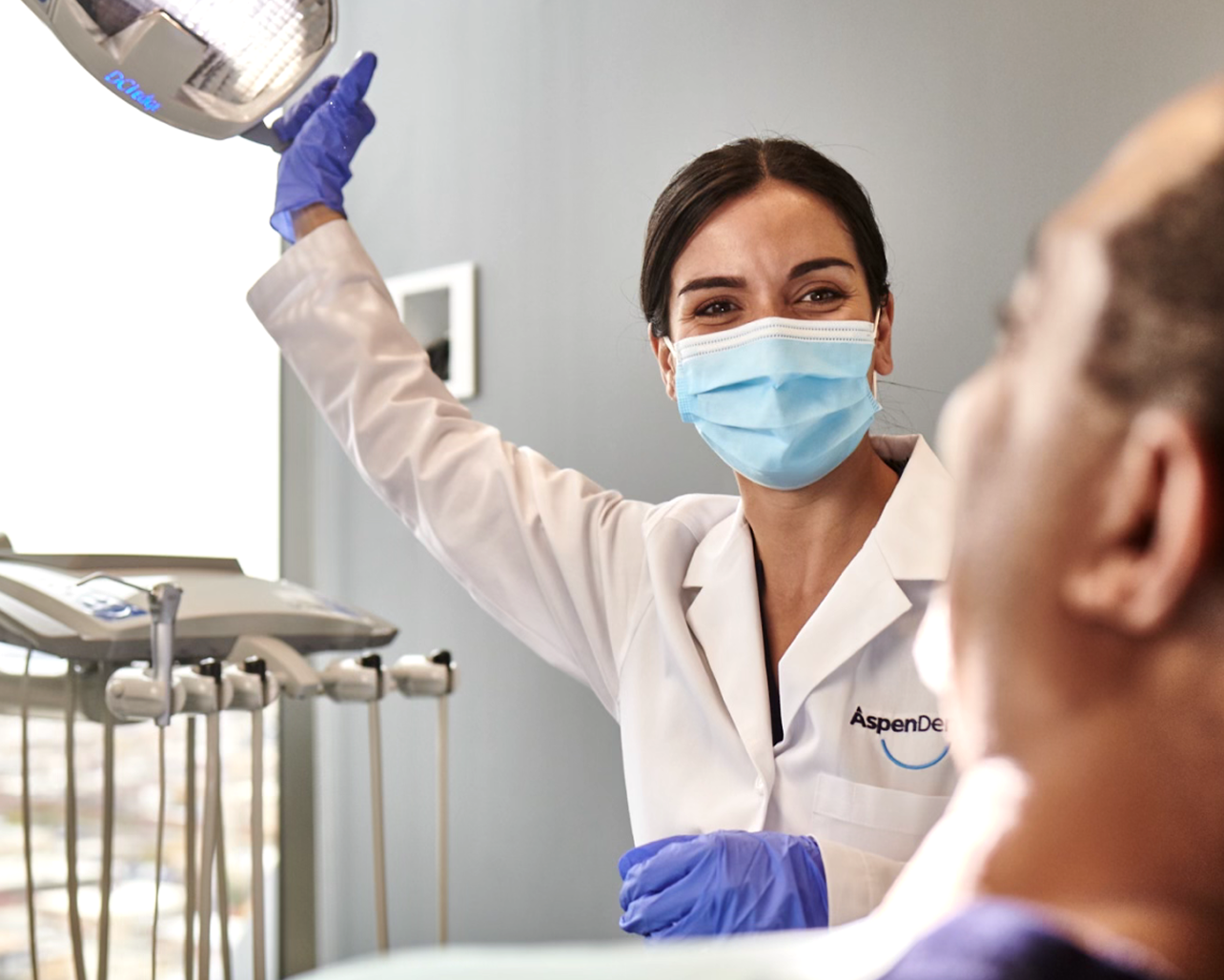 An Aspen Dental dentist wearing a medical mask and holding equipment speaks to a patient in a dental chair