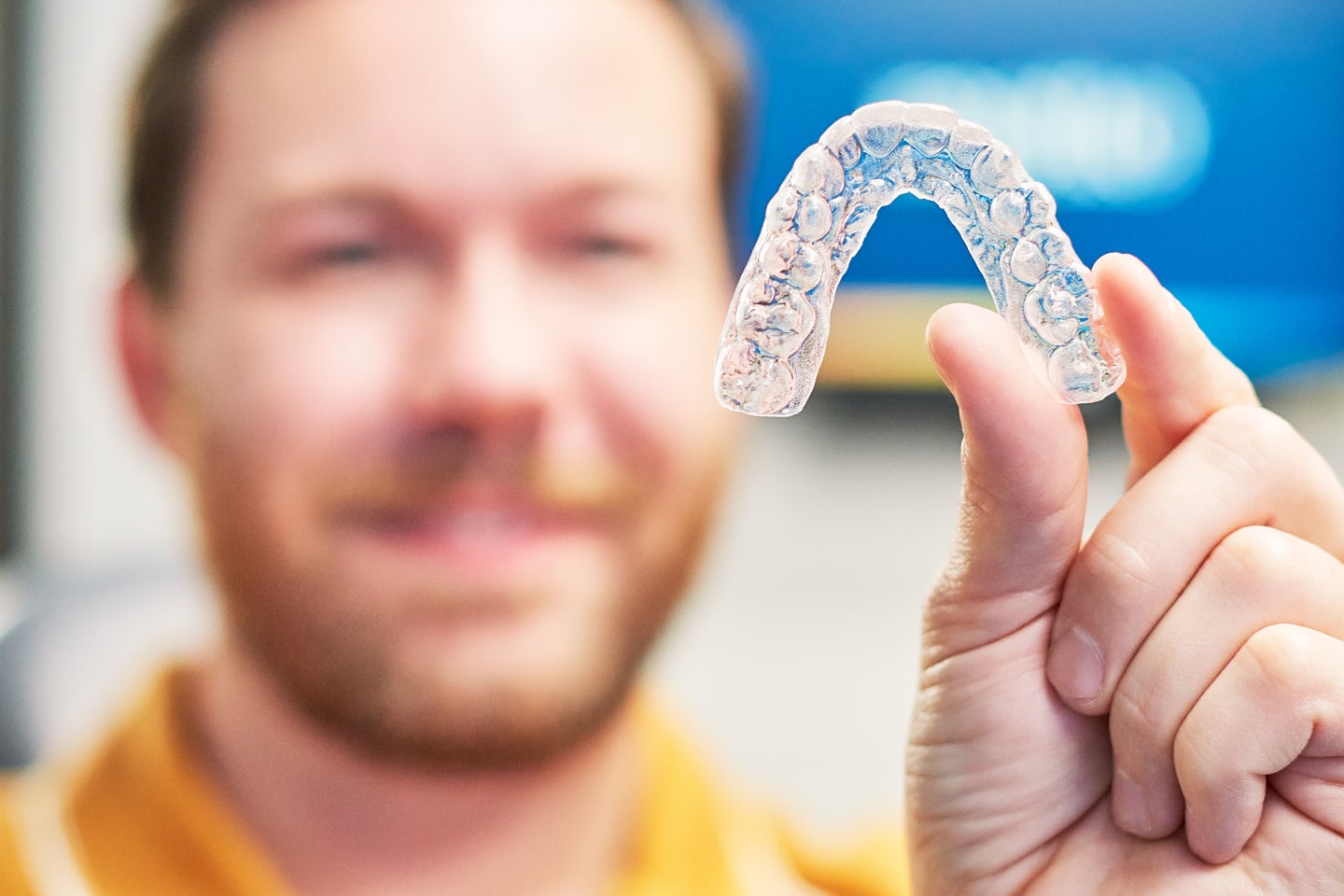 Smiling person holding Motto clear retainer close to the camera, showcasing its transparent design in a professional dental setting.