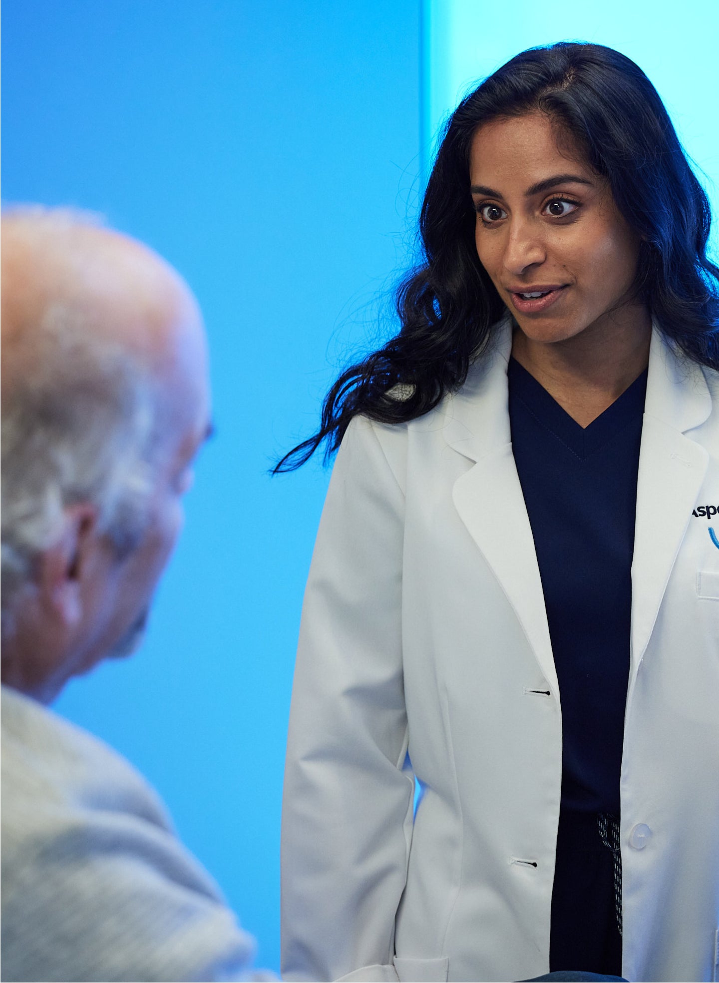 Aspen Dental professional wearing a white coat and navy scrubs engaging in a consultation with a patient in a modern clinical setting with a blue-toned background.
