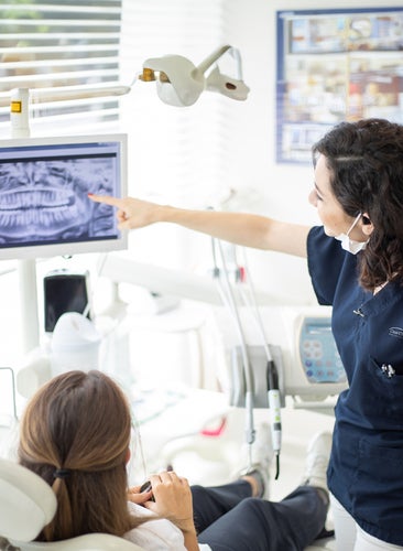 A dentist is talking to a patient in a dental chair while pointing at a screen.