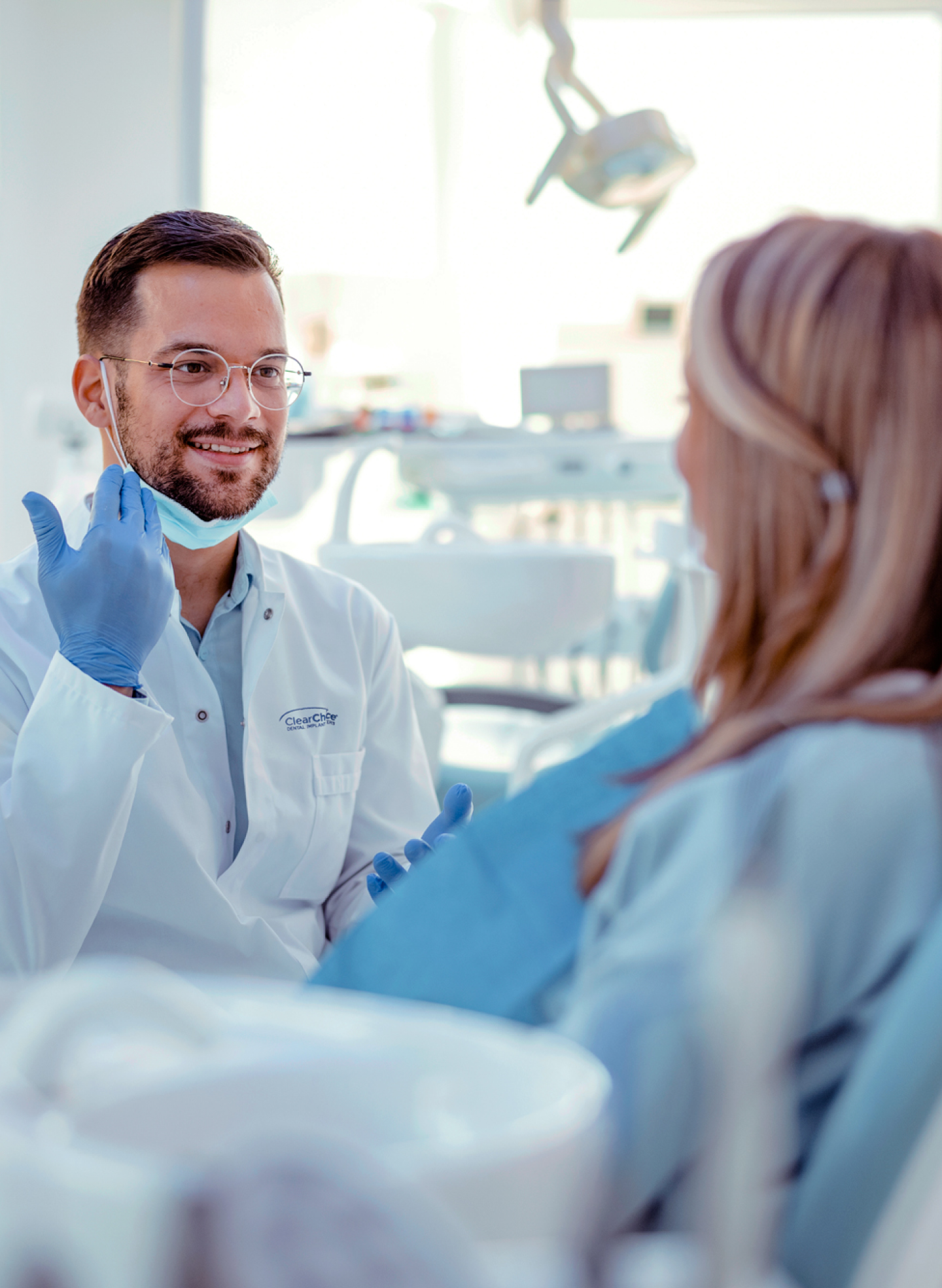 A ClearChoice dentist wearing gloves and a white coat discusses the dental implant recovery process with a patient in a modern dental office.