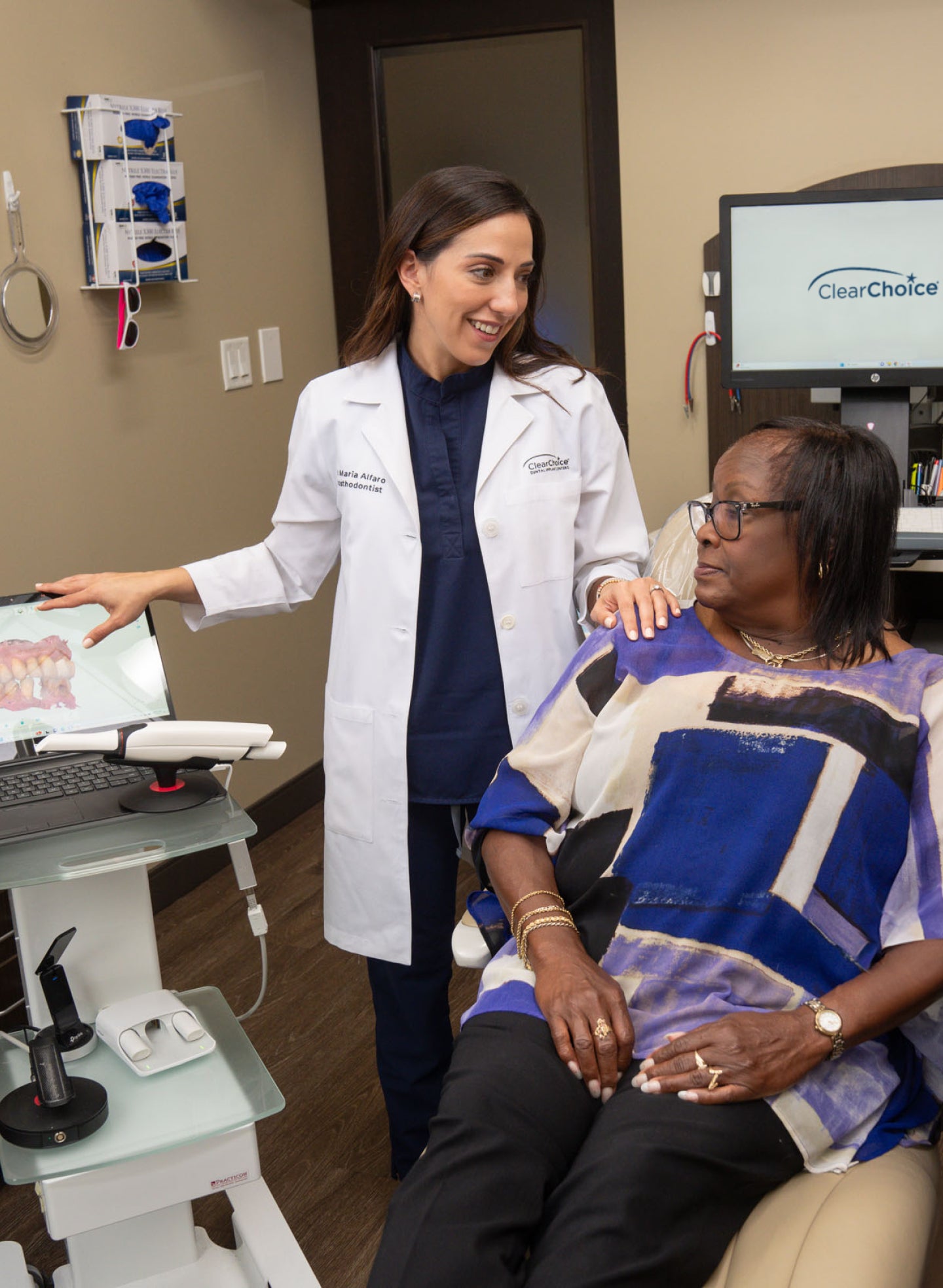 A ClearChoice dental specialist in a white lab coat explains osseointegration to a patient during a consultation. The doctor gestures towards a digital screen displaying a 3D model of teeth while the patient, seated in a dental chair, listens attentively. The ClearChoice logo is visible on a monitor in the background.