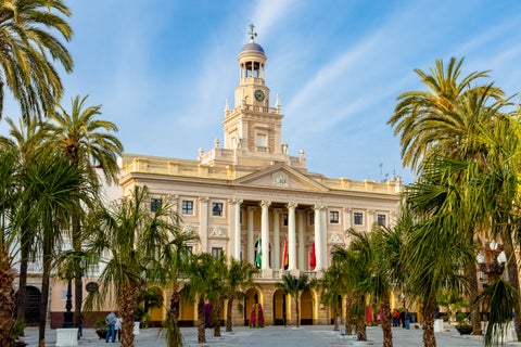 Cádiz Cathedral with a large cross atop its golden dome, framed by palm trees under a clear blue sky