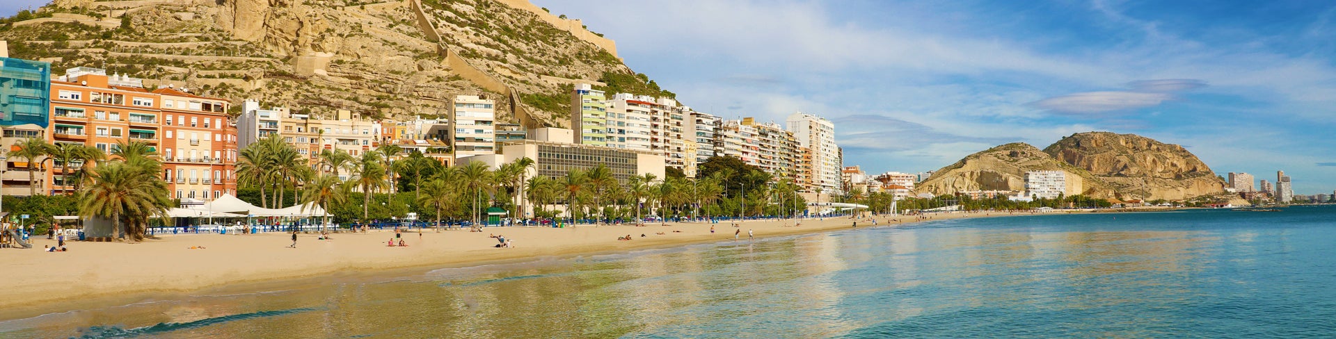 View of Alicante city and El Postiguet Beach on Spain's Costa Blanca, featuring golden sands, turquoise Mediterranean waters, palm-lined promenade, and Santa Bárbara Castle atop Mount Benacantil