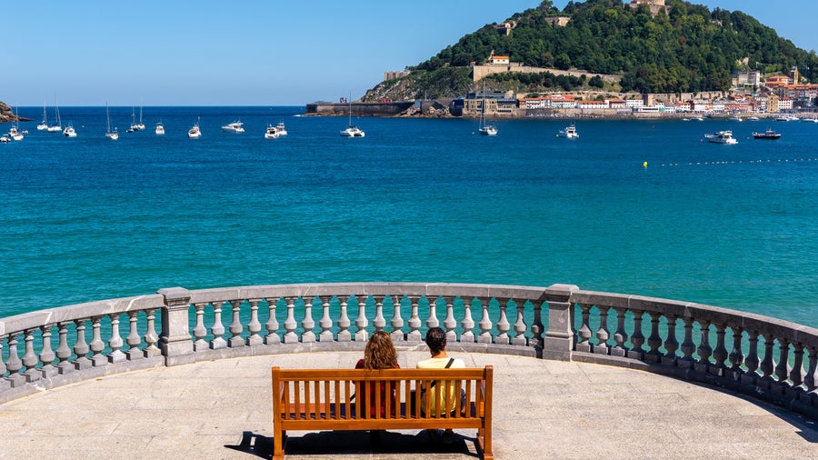 Tourists sitting on a bench overlooking the beach in San Sebastián, Spain, enjoying the seaside view