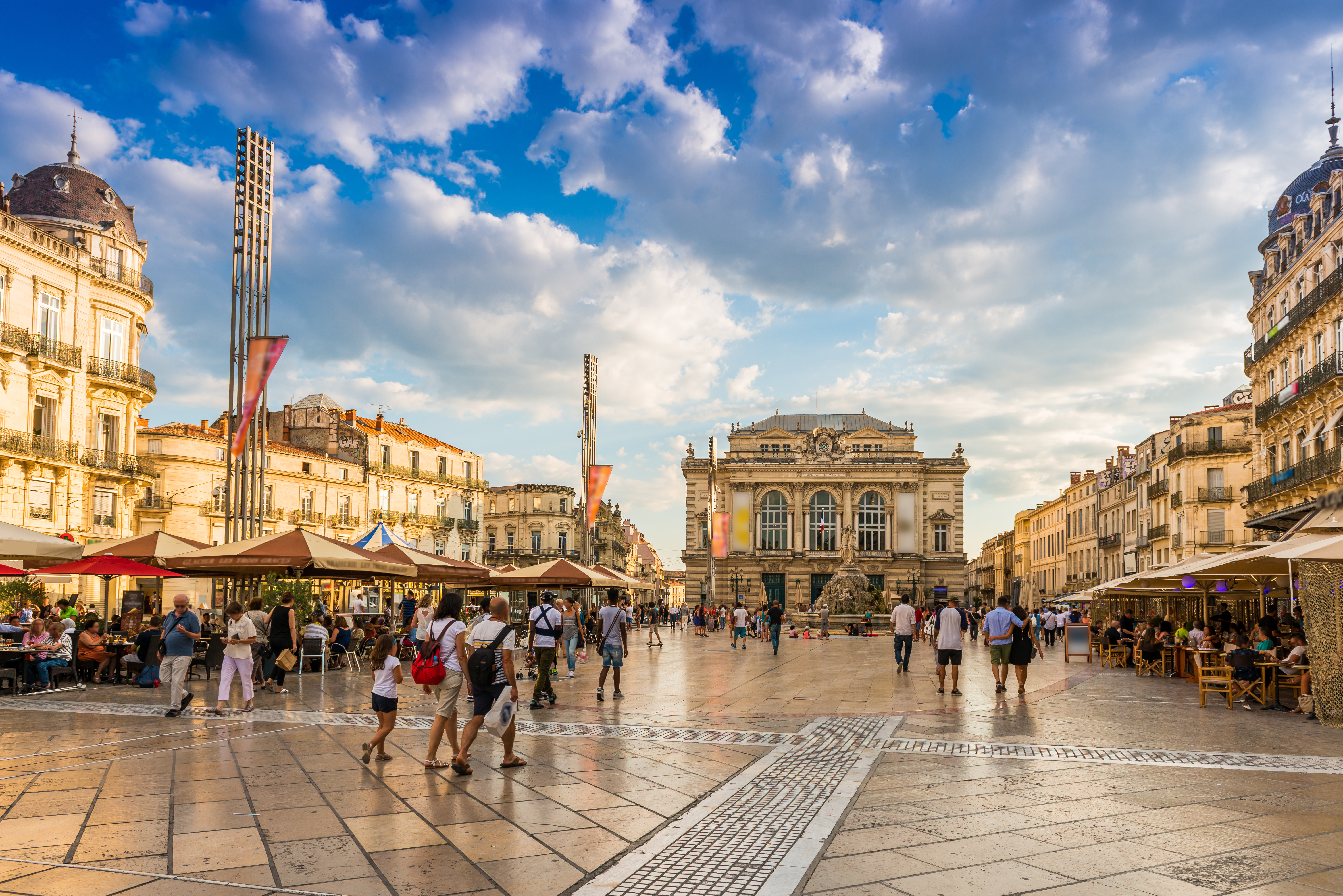 Place de la Comédie in Montpellier, France, with its vcentral square, framed by elegant Haussmann-style buildings, the iconic Opéra Comédie, and a lively atmosphere with pedestrians and outdoor café