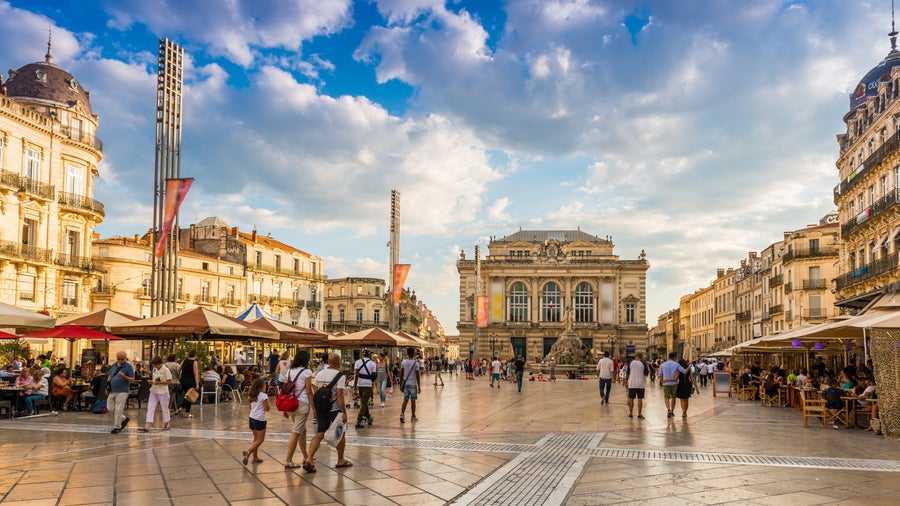 Place de la Comédie in Montpellier, France, with its vcentral square, framed by elegant Haussmann-style buildings, the iconic Opéra Comédie, and a lively atmosphere with pedestrians and outdoor café