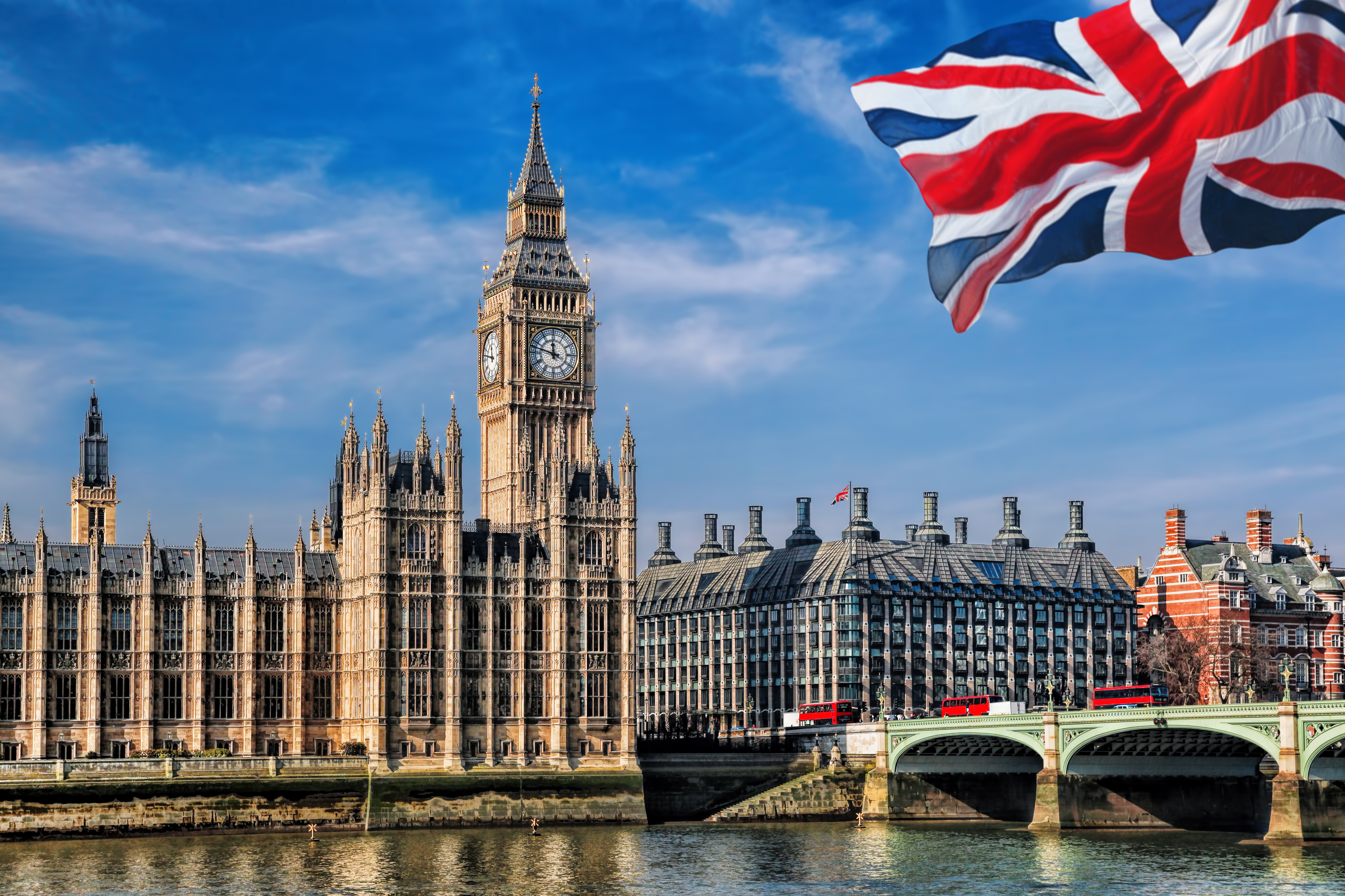 Big Ben and Westminster with Westminster Bridge and red buses crossing, viewed from across the River Thames with UK flag