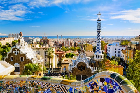 Sunny view of Park Güell in Barcelona, Spain, showing vibrant mosaic tiles, playful architectural elements, and green gardens with the city in the distance