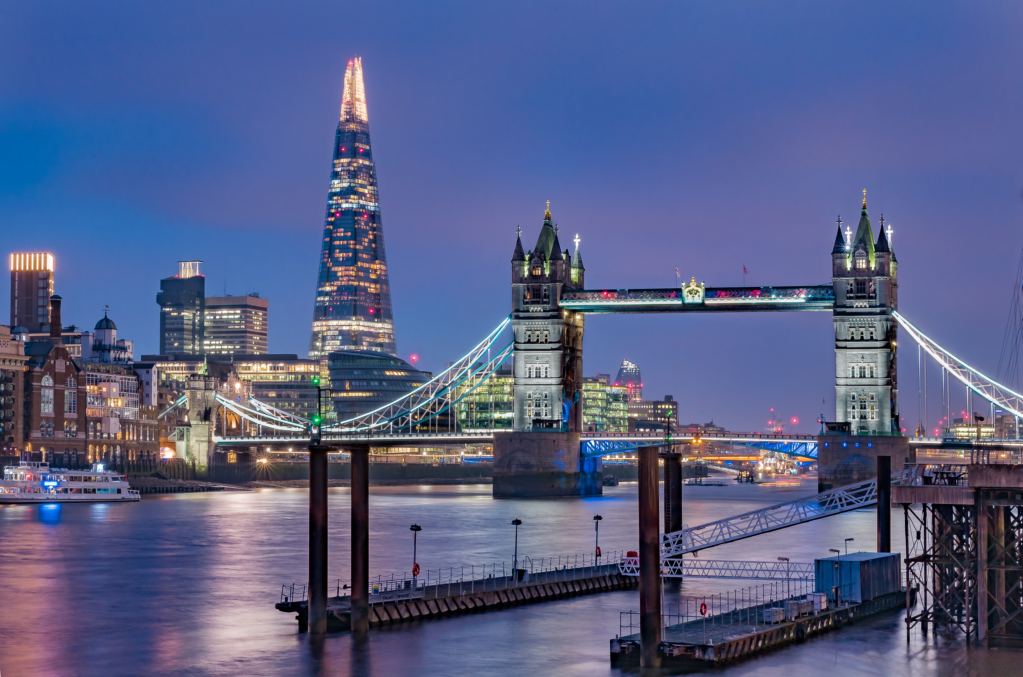 Tower Bridge illuminated at night with modern London buildings in the background, viewed from the River Thames