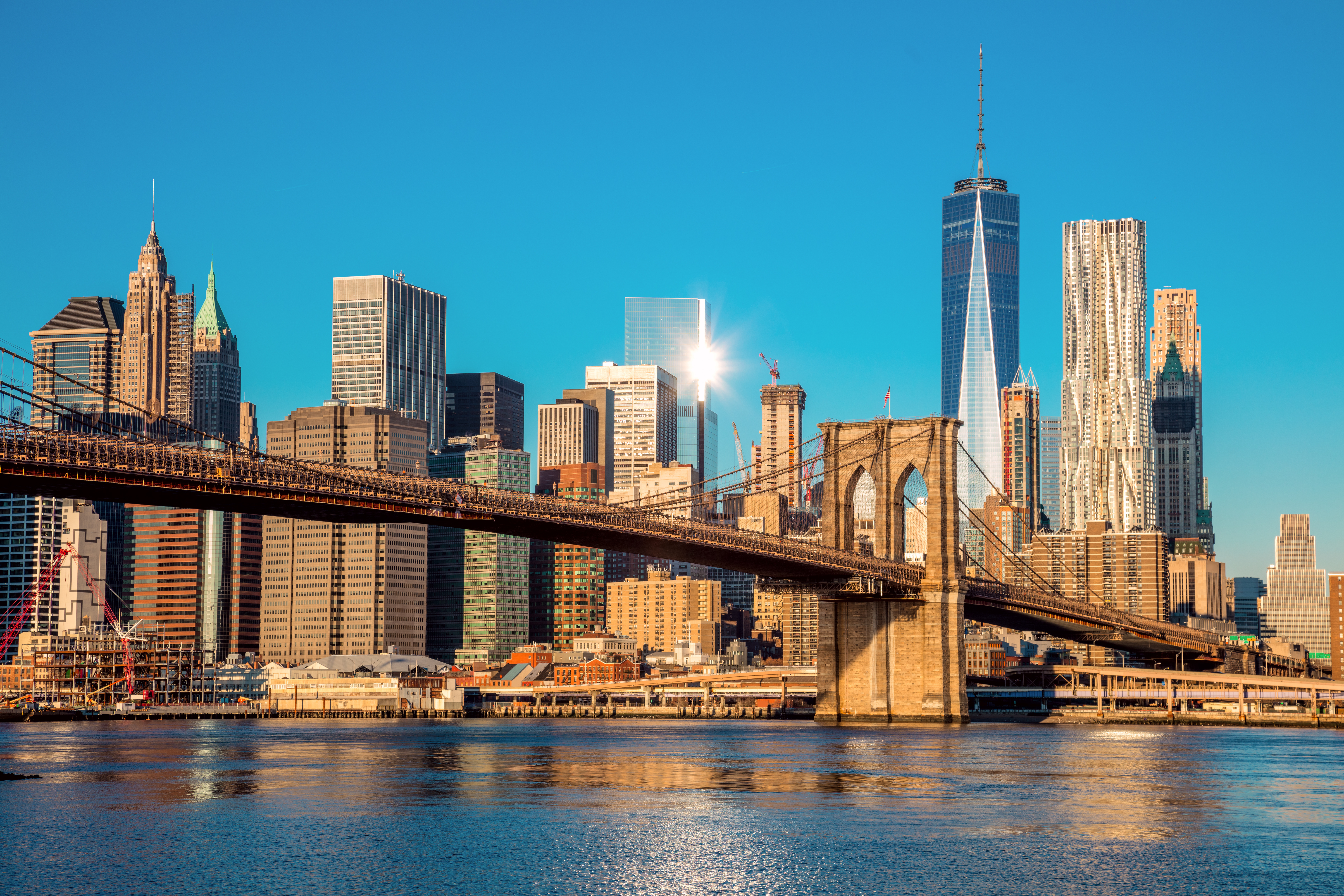 Brooklyn Bridge from below over East River with Manhattan skyline at sunrise and blue sky, New York City