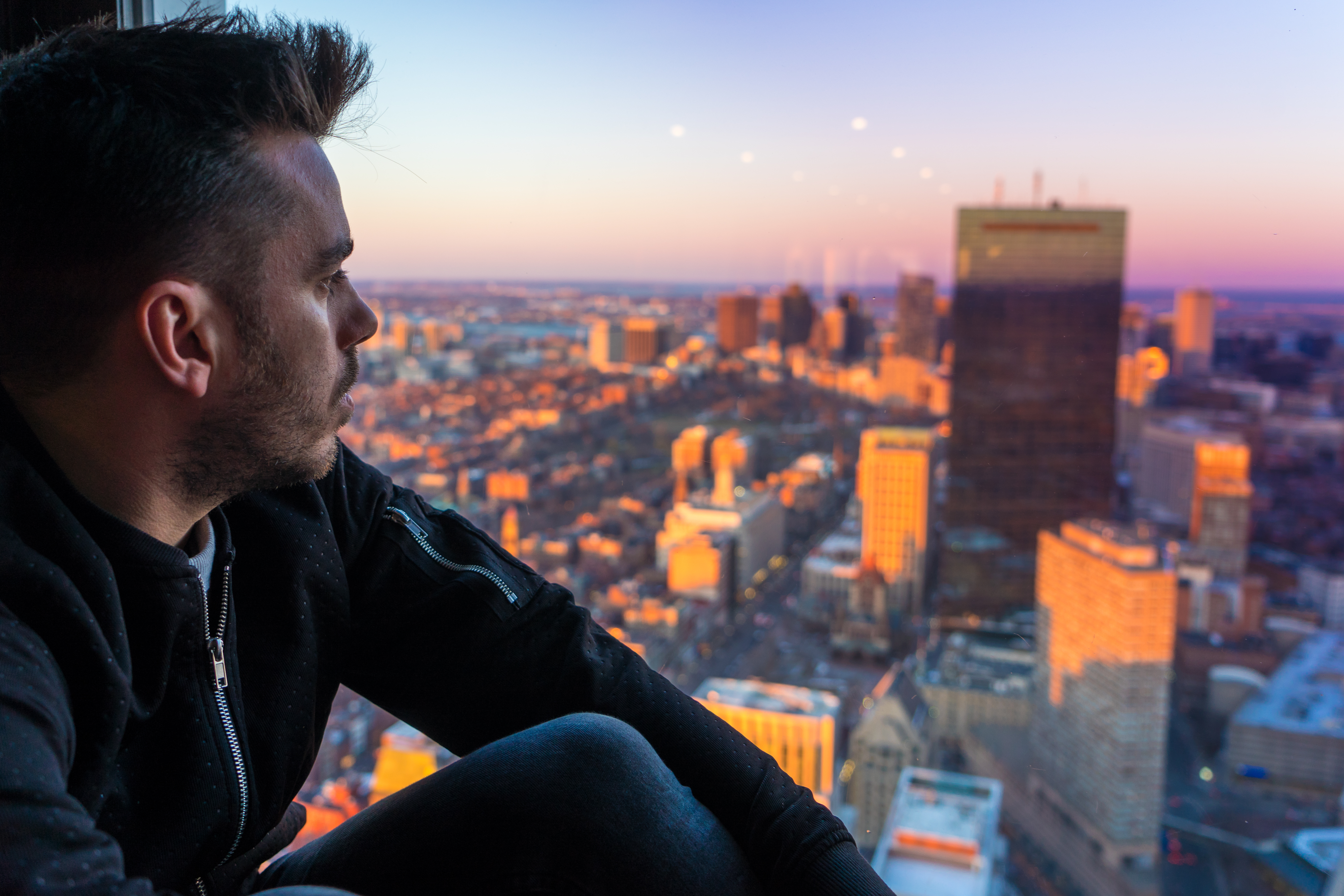 Man student sitting by high-rise window overlooking Boston skyline at pink sunset