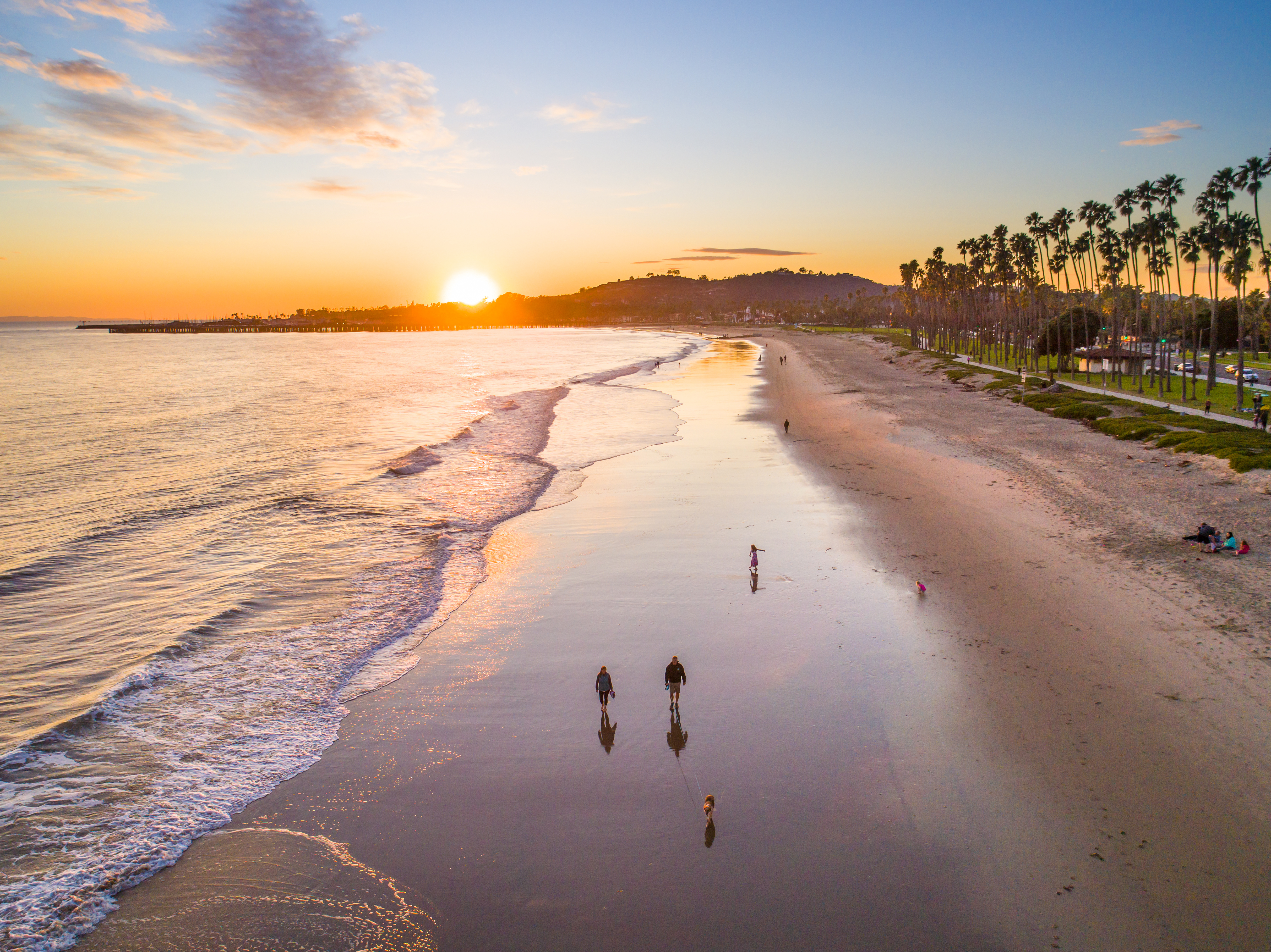 Aerial view of East Beach, Santa Barbara, California at sunset with white sand, palm trees, and family walking with children and dog
