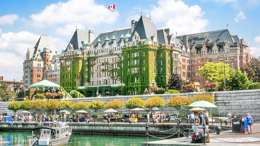 Sailboats and small ferries float on the calm, reflective waters, surrounded by the vibrant architecture of historic buildings and modern establishments in Inner Harbour in Victoria, British Columbia, Canada