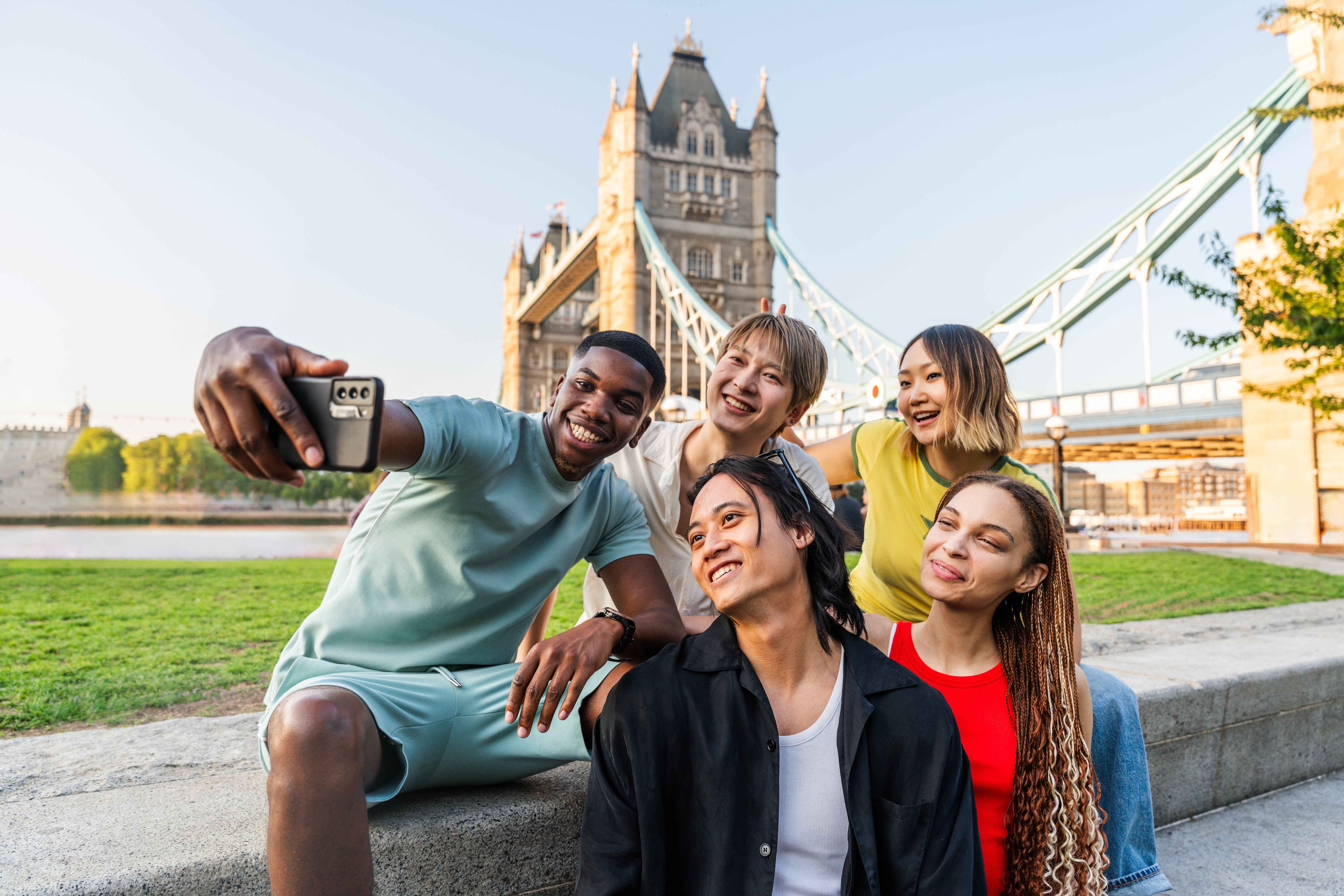 Group of young international students taking a selfie on the riverside grass with Tower Bridge in London in summer